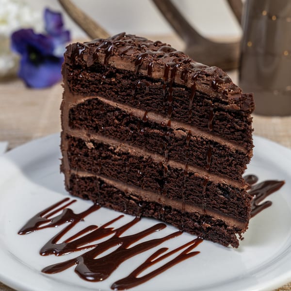 A rich slice of chocolate cake with chocolate frosting and drizzle on a white plate.