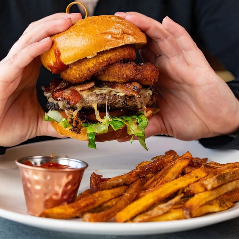 Hands holding a juicy burger with bacon, cheese, and onion rings, served with fries and ketchup.