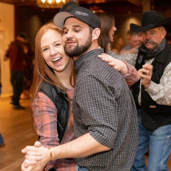 A smiling couple dances closely, with other people and a man in a cowboy hat in the background.